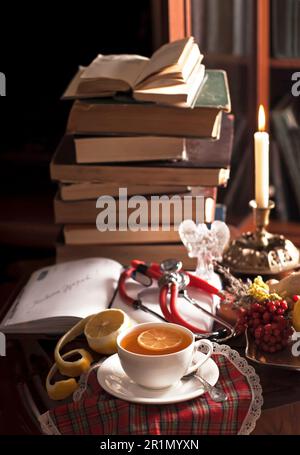 Une pile de livres anciens, un phonendoscope, une tasse de thé au citron, un bloc-notes ouvert, des plantes médicinales sur une table en bois Banque D'Images