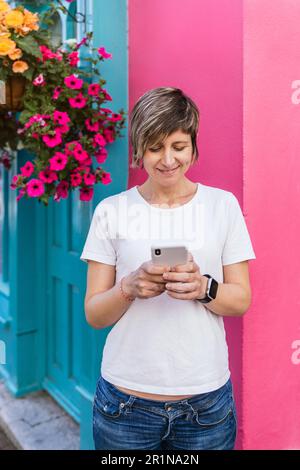 Femme blonde souriante, captivée par le charme irlandais dynamique, la messagerie texte au milieu de murs colorés et de délices floraux Banque D'Images