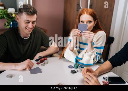 Drôle redhead femme jeune femme fait duper tenant des cartes par le visage pendant la fête d'amusement à la maison le week-end jouant au jeu de poker avec des amis. Banque D'Images