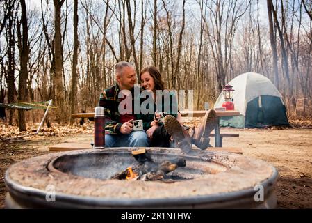 Un couple souriant se détend par un feu de camp en buvant du café à l'automne Banque D'Images