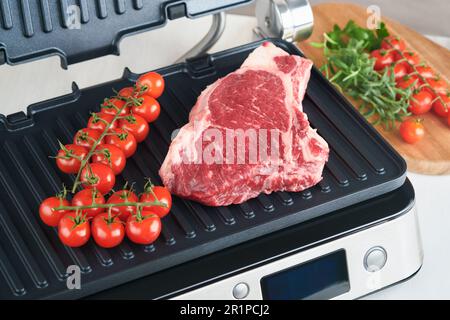 Steaks de première qualité crus sur le gril électrique. Steaks de viande de choix black angus Ribeye sur un grill électrique moderne au romarin, à la tomate cerise et au poivre. Cuisine pro Banque D'Images