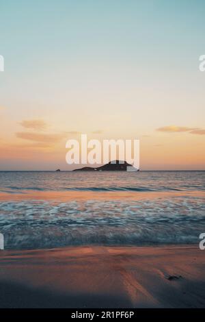 Paysage vertical de lever de soleil sur l'océan avec île et ciel coloré en arrière-plan. Vagues et rives sur l'eau de mer sous la lumière du matin. Espace de copie du ciel vide. Beauté nature paysage marin au coucher du soleil moody soir crépuscule. Banque D'Images