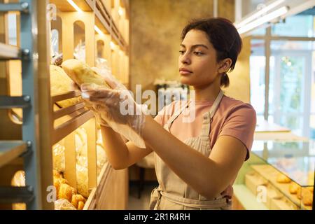 Jeune femme sérieuse en tablier de coton beige prenant du pain frais exposé tout en servant les clients de la boulangerie ou de la cafétéria Banque D'Images