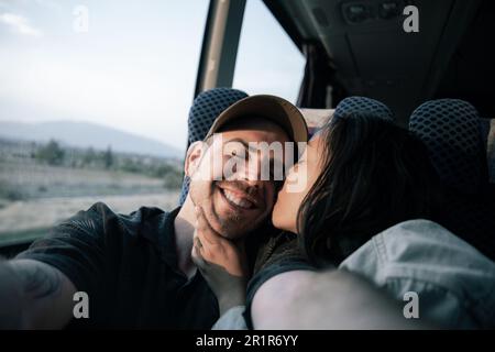 Un jeune couple hispanique et caucasien prend un selfie avec un baiser sur la joue pendant un voyage en bus Banque D'Images