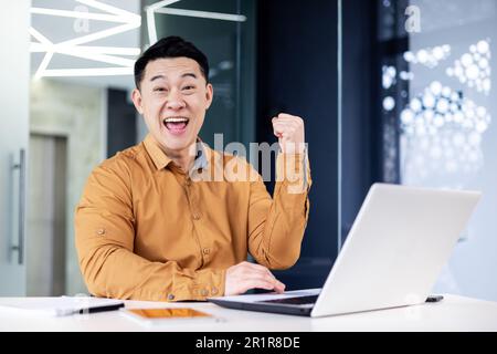 Homme d'affaires asiatique couronné de succès, portrait de l'homme au bureau en chemise décontractée, employé souriant et regardant l'appareil photo satisfait de la réussite en tenant le geste de la victoire main vers le haut Banque D'Images