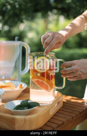 Femme préparant une carafe d'eau infusée avec des fruits frais et des feuilles de menthe dans le jardin Banque D'Images