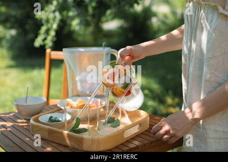 Femme versant un verre d'eau avec un assortiment de fruits frais et de feuilles de menthe dans le jardin Banque D'Images