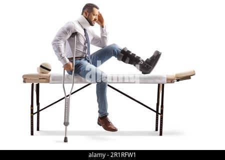 Homme blessé avec une chaussure orthopédique et un col cervical assis sur une table de thérapie et tenant sa tête isolée sur fond blanc Banque D'Images