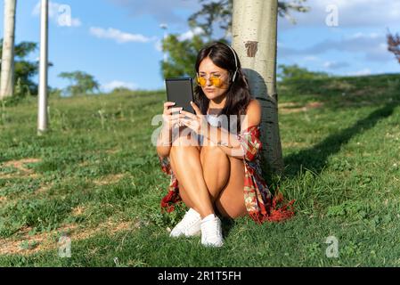 Femme avec des lunettes de soleil et des écouteurs appréciant la lecture d'un ebook tout en se relaxant assis sur l'herbe dans un parc. Concept de technologie. Banque D'Images