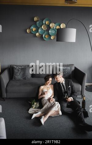 mariée gaie en robe de mariage et marié en costume noir buvant du champagne tout en célébrant leur mariage près du bouquet de mariée, canapé et lampe de plancher Banque D'Images