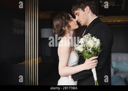 vue latérale d'un beau marié en noir habillé porter embrasser brunette mariée dans l'élégante robe de mariage tenant le bouquet de mariée avec des fleurs tout en se tenant Banque D'Images