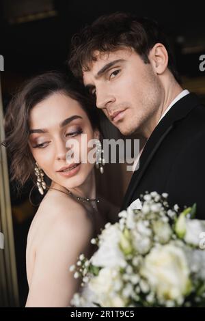bonne tenue élégante en noir tenue habillée debout avec brunette et mariée élégante avec maquillage et bijoux tenant le bouquet de mariée tout en se tenant dans le fourre-tout Banque D'Images