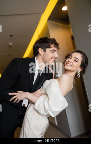 groom gai en costume noir se penchant vers la charmante mariée en robe de mariage blanche tout en souriant ensemble et debout dans le hall de l'hôtel moderne, couple Banque D'Images