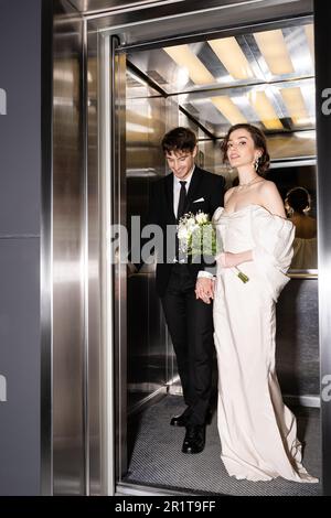 pleine longueur de jolie mariée en robe blanche tenant le bouquet de mariée avec des fleurs et main de groom gai gai gai en costume debout dans l'ascenseur dans l'hôtel, heureux Banque D'Images