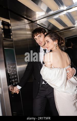 très beau marié en tenue formelle en appuyant sur bouton tout en embrassant la mariée de brunette dans la robe de mariage et debout ensemble dans l'ascenseur de l'hôtel moderne, stock Banque D'Images