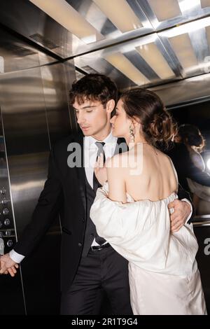 beau marié en tenue formelle en appuyant sur bouton tout en embrassant brunette et jeune mariée dans la robe de mariage et debout ensemble dans ascenseur de l'h moderne Banque D'Images