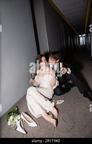 joyeux marié en costume noir tenant une bouteille et assis près de la superbe mariée avec un verre de champagne à côté du bouquet de mariée et des talons hauts sur le sol en cor Banque D'Images