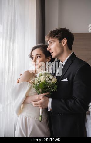 superbe groom dans une tenue classique debout avec une bouche ouverte à côté de l'élégante jeune mariée en bijoux, robe blanche avec bouquet de mariée dans un hôtel moderne Banque D'Images