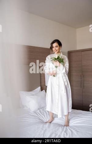 jeune mariée gaie avec cheveux de brunette en robe de soie blanche et boucles d'oreilles de perle tenant le bouquet de mariée tout en se tenant sur le lit dans la chambre d'hôtel, occa spécial Banque D'Images