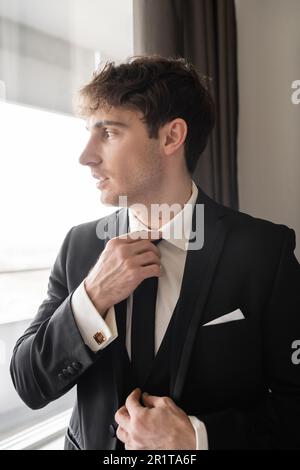 très beau dans un élégant vêtement habillé avec chemise blanche réglable cravate noire tout en regardant la fenêtre dans une chambre d'hôtel moderne, homme le jour du mariage, prepar Banque D'Images