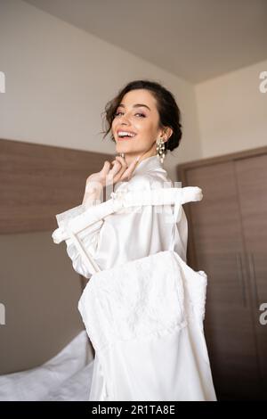 mariée joyeuse avec cheveux de brunette en robe de soie blanche tenant le crochet doux avec élégante robe de mariage et souriant dans la chambre d'hôtel, occasions spéciales Banque D'Images