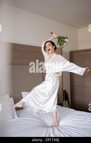 jeune mariée excitée avec cheveux de brunette en robe de soie blanche et boucles d'oreilles de perle tenant le bouquet de mariée tout en sautant sur le lit dans la chambre d'hôtel, occasion spéciale Banque D'Images