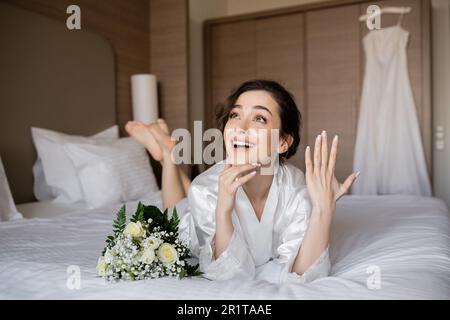 femme heureuse avec cheveux brunette, robe de soie blanche couchée et montrant l'anneau d'engagement sur le doigt à côté du bouquet de mariée sur le lit dans la chambre d'hôtel avec le dr de mariage Banque D'Images