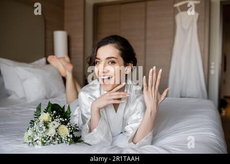 femme stupéfait avec des cheveux brunette, robe de soie blanche couchée et montrant l'anneau d'engagement sur le doigt à côté du bouquet de mariée sur le lit dans la chambre d'hôtel avec mariage d Banque D'Images