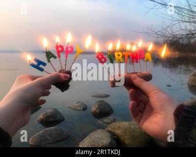 Inscription d'anniversaire brûlante faite de bougies de vacances dans les mains d'un homme et d'une femme face à l'eau du lac de l'océan. Concept: birt Banque D'Images