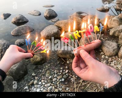 Inscription d'anniversaire brûlante faite de bougies de vacances dans les mains d'un homme et d'une femme face à l'eau du lac de l'océan. Concept: birt Banque D'Images