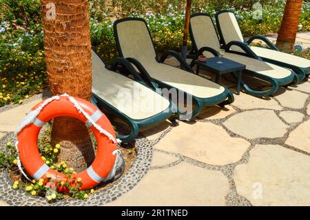 La bouée de sauvetage orange pour la sécurité de la baignade repose sur un sol en pierre sur un fond de fleurs jaunes et de chaises longues par l'exoti marin tropical Banque D'Images