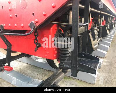 Grandes roues en fer d'un train rouge et noir debout sur des rails et des éléments de suspension avec ressorts d'une ancienne locomotive industrielle à vapeur. Banque D'Images