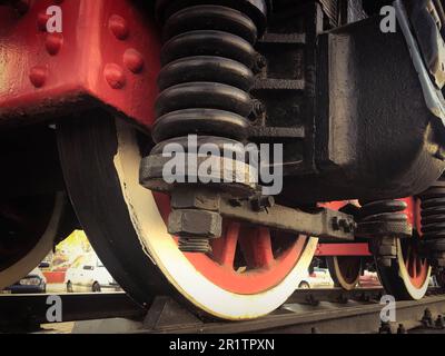 Grandes roues en fer d'un train rouge et noir debout sur des rails et des éléments de suspension avec ressorts d'une ancienne locomotive industrielle à vapeur. Banque D'Images