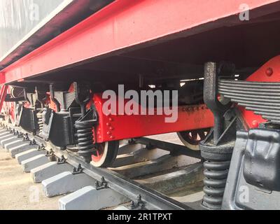 Grandes roues en fer d'un train rouge et noir debout sur des rails et des éléments de suspension avec ressorts d'une ancienne locomotive industrielle à vapeur. Banque D'Images