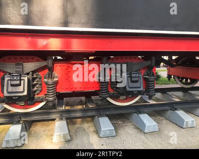 Grandes roues en fer d'un train rouge et noir debout sur des rails et des éléments de suspension avec ressorts d'une ancienne locomotive industrielle à vapeur. Banque D'Images