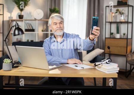 Homme souriant environ 65 posant avec le doigt pointant devant l'appareil photo de smartphone tout en prenant une pause de travail dans le bureau à domicile. Travailleur indépendant mature prenant le selfie de l'image confortable de style de vie en intérieur. Banque D'Images