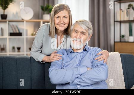 Portrait de l'homme de famille aimant et de la femme en tenue décontractée passer du temps ensemble dans un studio moderne. Une dame gaie d'âge moyen qui embrasse un homme chaleureusement heureux tout en se tenant derrière un canapé dans le salon. Banque D'Images