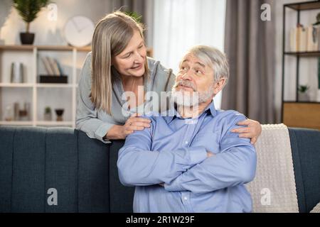 Portrait de l'homme de famille aimant et de la femme en tenue décontractée passer du temps ensemble dans un studio moderne. Une dame gaie d'âge moyen qui embrasse un homme chaleureusement heureux tout en se tenant derrière un canapé dans le salon. Banque D'Images