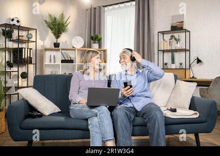 Homme mûr gai utilisant des écouteurs sans fil et mobile tandis que belle femme d'âge moyen se détendant avec un ordinateur portable dans le salon. Les conjoints seniors écoutent de la musique d'excellente qualité dans une liste de lecture moderne à la maison. Banque D'Images