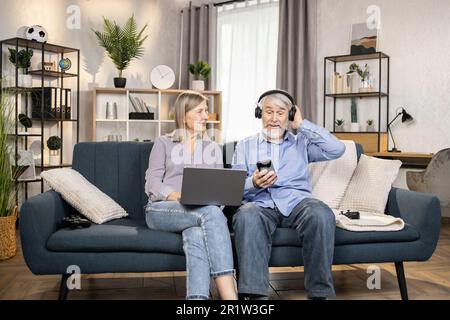 Homme mûr gai utilisant des écouteurs sans fil et mobile tandis que belle femme d'âge moyen se détendant avec un ordinateur portable dans le salon. Les conjoints seniors écoutent de la musique d'excellente qualité dans une liste de lecture moderne à la maison. Banque D'Images