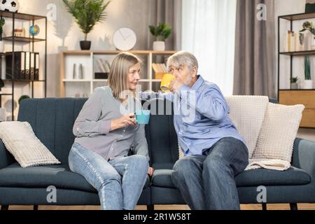 Un couple caucasien à la retraite appréciant un délicieux thé chaud tout en discutant les uns avec les autres dans une chambre confortable et élégante. Un mari et une femme heureux se reposant sur un canapé confortable discutant de plans passionnants pour le week-end. Banque D'Images