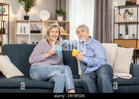 Un couple caucasien à la retraite appréciant un délicieux thé chaud tout en discutant les uns avec les autres dans une chambre confortable et élégante. Un mari et une femme heureux se reposant sur un canapé confortable discutant de plans passionnants pour le week-end. Banque D'Images