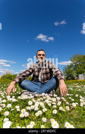 Un jeune homme beau est assis et s'amuser dans un pré fleuri ensoleillé par une journée ensoleillée. Jeune homme détendu assis dans un champ de fleurs Banque D'Images