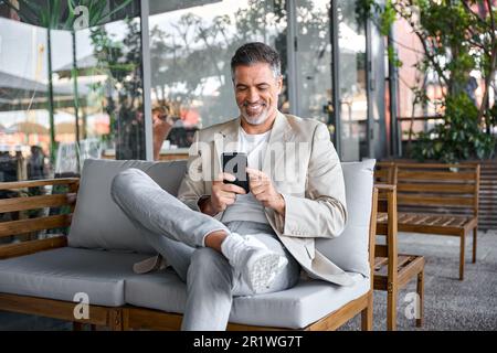 Joyeux homme d'affaires décontracté d'un âge moyen assis dans un café extérieur à l'aide d'un smartphone. Banque D'Images