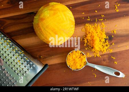 Cuillère à soupe de zeste d'orange frais sur une planche à découper en bois : une orange à zestes avec une boîte de râpe vue directement au-dessus Banque D'Images