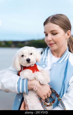Vue d'une femme blonde portant son chien maltais à fourrure blanche, lac à l'arrière-plan Banque D'Images