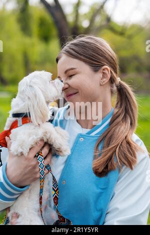 Vue d'une femme blonde tenant son chien maltais avec de la fourrure blanche alors qu'il est le nez de propriétaire de licence, la nature sur l'arrière-plan Banque D'Images