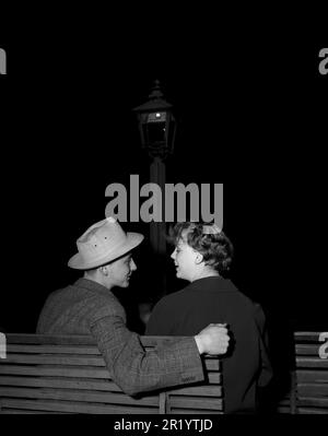 Dans le 1950s. Un jeune couple est assis sur un banc et il regarde af il fait une tentative de mettre son bras autour d'elle, le prenant pas à pas. Une première date peut-être. Suède 1953 Kristoffersson réf. 15K-21 Banque D'Images