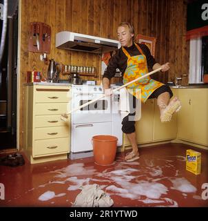 Jour de nettoyage en 1970s. Intérieur d'une cuisine où l'on voit un homme nettoyer le sol. Pour accélérer le travail, il a monté des brosses sur ses pieds. Il est acteur Stig Grybe 1928-2017. 4 octobre 1972. Kristoffersson Banque D'Images