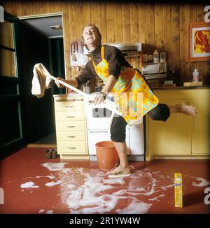 Jour de nettoyage en 1970s. Intérieur d'une cuisine où l'on voit un homme nettoyer le sol. Pour accélérer le travail, il a monté des brosses sur ses pieds. Il est acteur Stig Grybe 1928-2017. 4 octobre 1972. Kristoffersson Banque D'Images